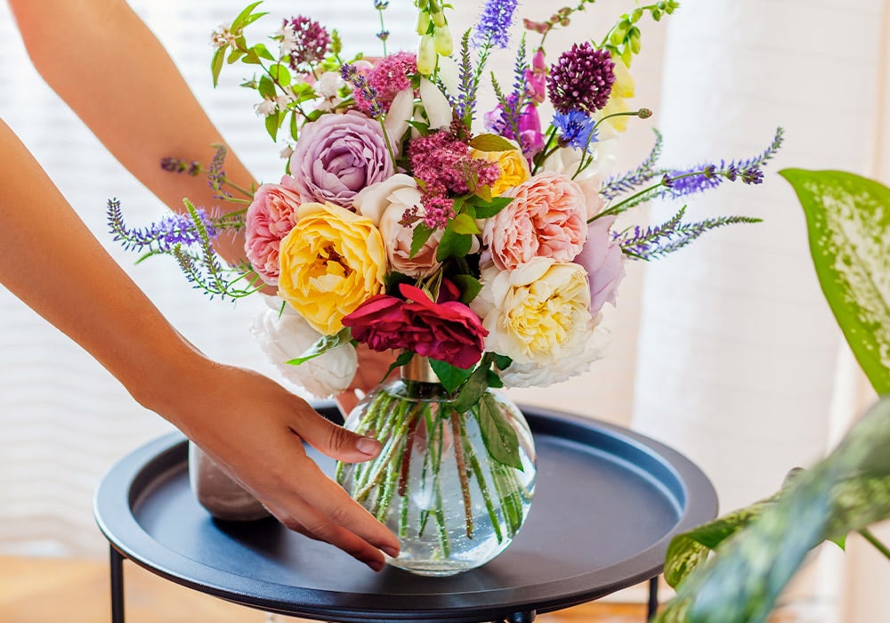 A person places a vibrant, multi-colored flower bouquet in a clear round vase on a small, circular black table in a brightly lit room.